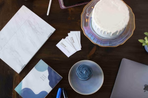 A desk setup with a laptop, a blue notebook, and a white cake, featuring CAYK Marketing business cards to illustrate the synergy between branding vs. marketing.