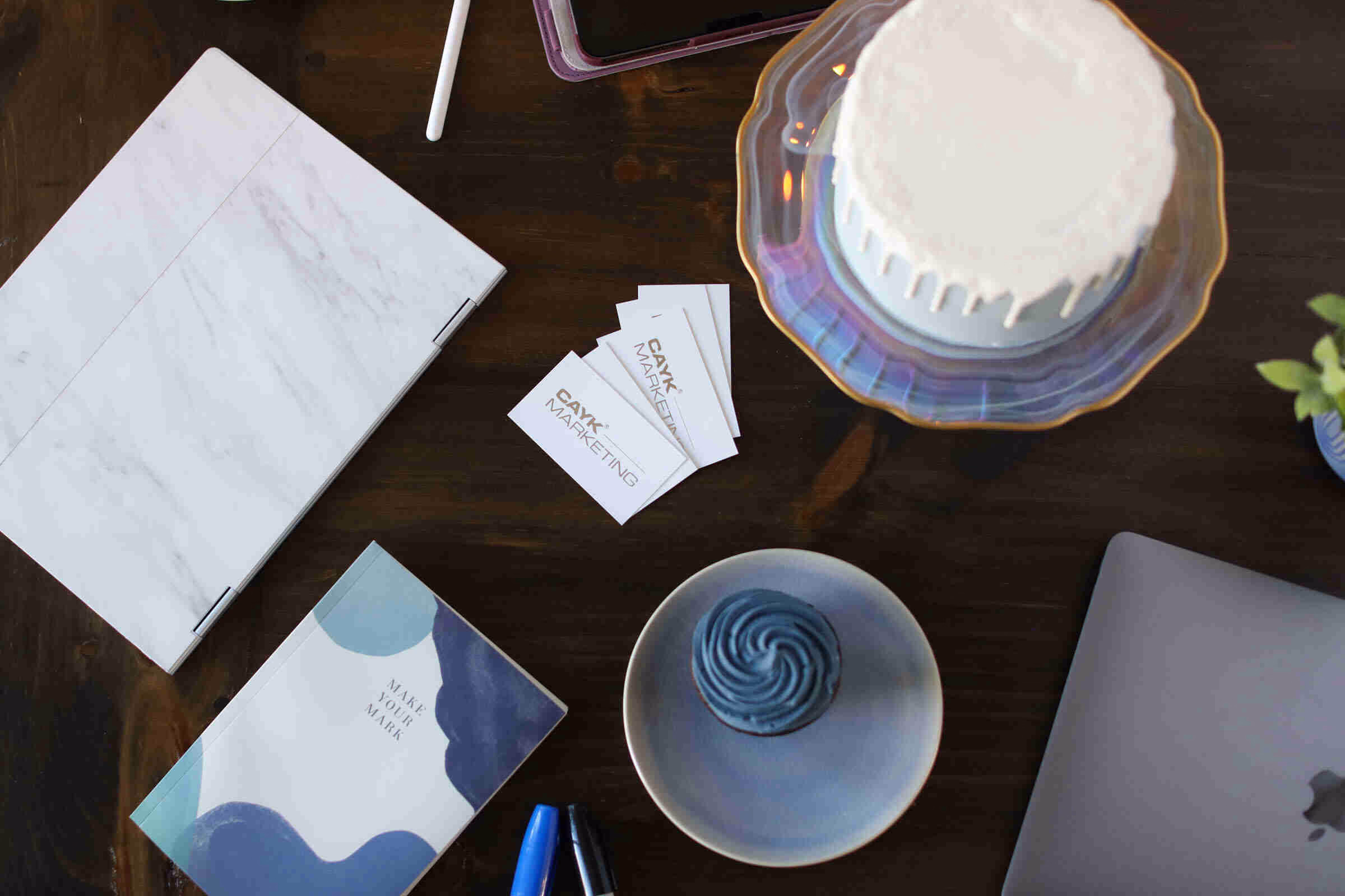 A desk setup with a laptop, a blue notebook, and a white cake, featuring CAYK Marketing business cards to illustrate the synergy between branding vs. marketing.