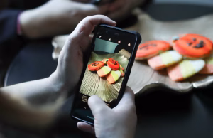 Person capturing pumpkin and candy corn cookies with a phone for Halloween social media campaigns.